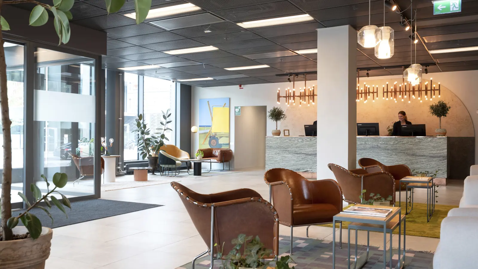 Modern hotel lobby with stylish brown leather chairs, plants, and a reception desk in the background. The area has contemporary lighting and decor, creating a welcoming atmosphere.