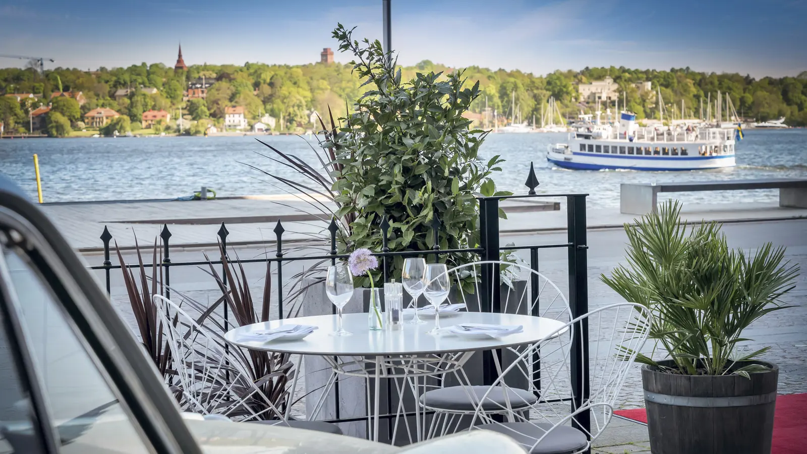 Riverside outdoor dining setup with a white table, wine glasses, and chairs, surrounded by greenery, overlooking a scenic waterfront with boats and lush trees in the distance.