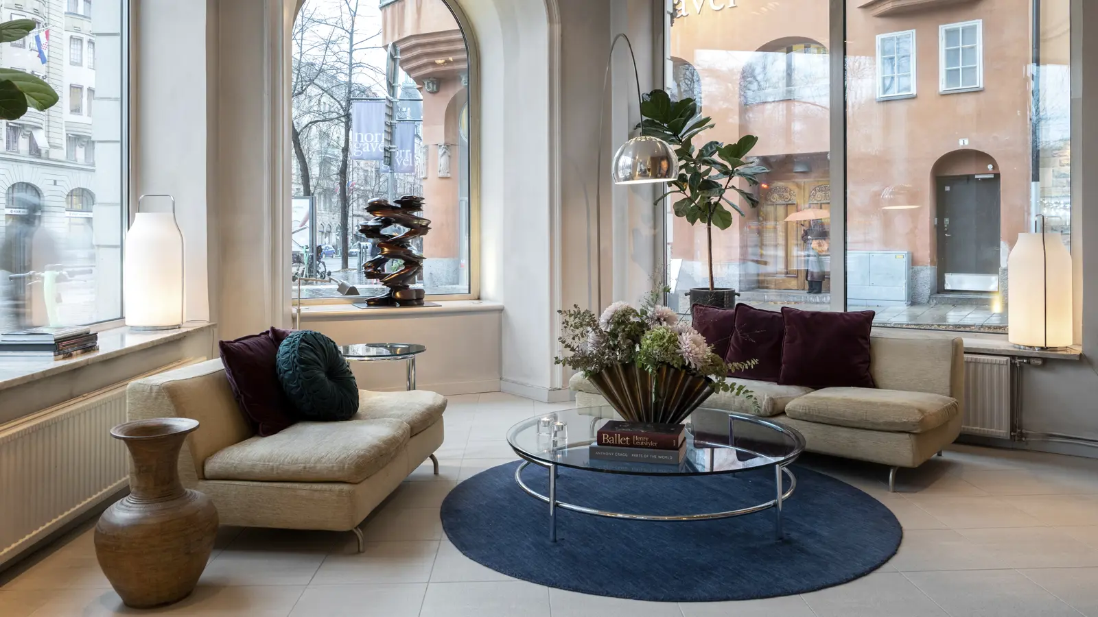 Modern lounge area with beige sofas, large windows, and elegant decor, featuring a round glass coffee table, blue rug, and indoor plants, set against an urban backdrop.