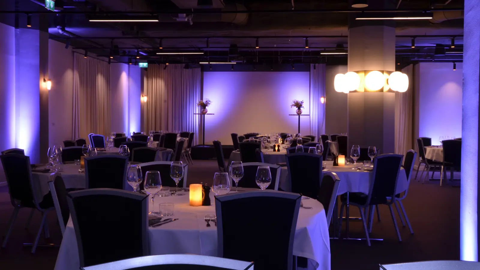 Elegant banquet hall with round tables, wine glasses, and candles, illuminated by soft purple lighting and modern chandeliers.