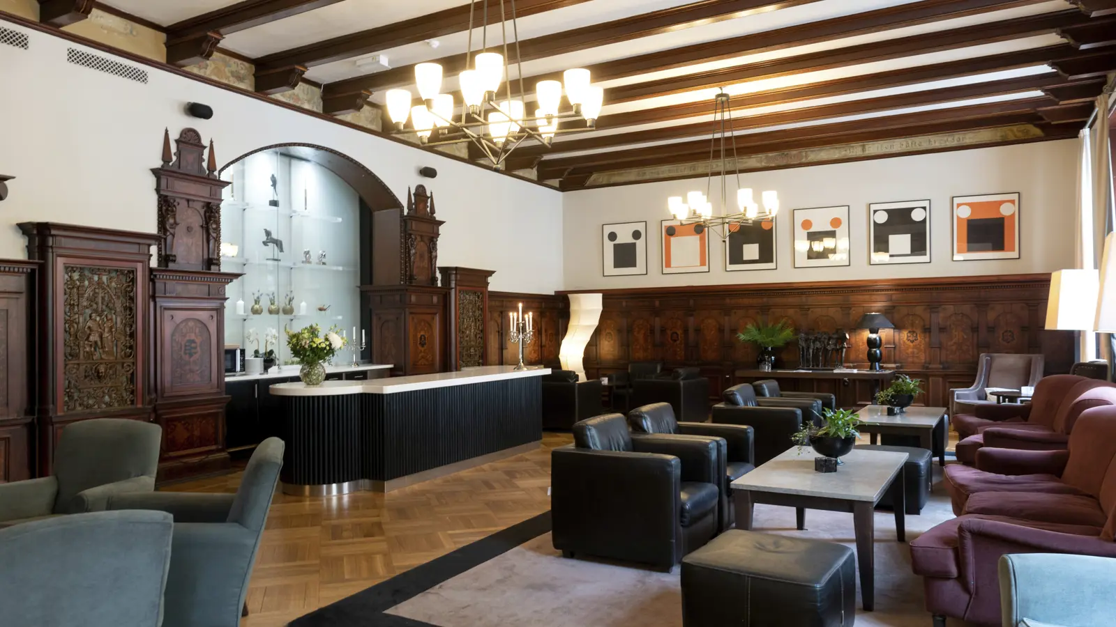 Elegant hotel lounge with dark leather chairs, art deco chandeliers, and decorative wood paneling, featuring a central modern bar and contemporary artwork on the walls.