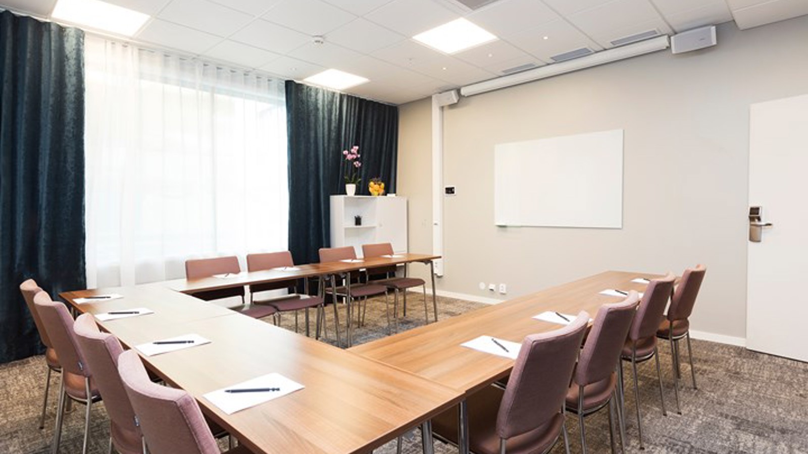 Conference room with U-shaped seating, wooden tables, light walls, dark curtains