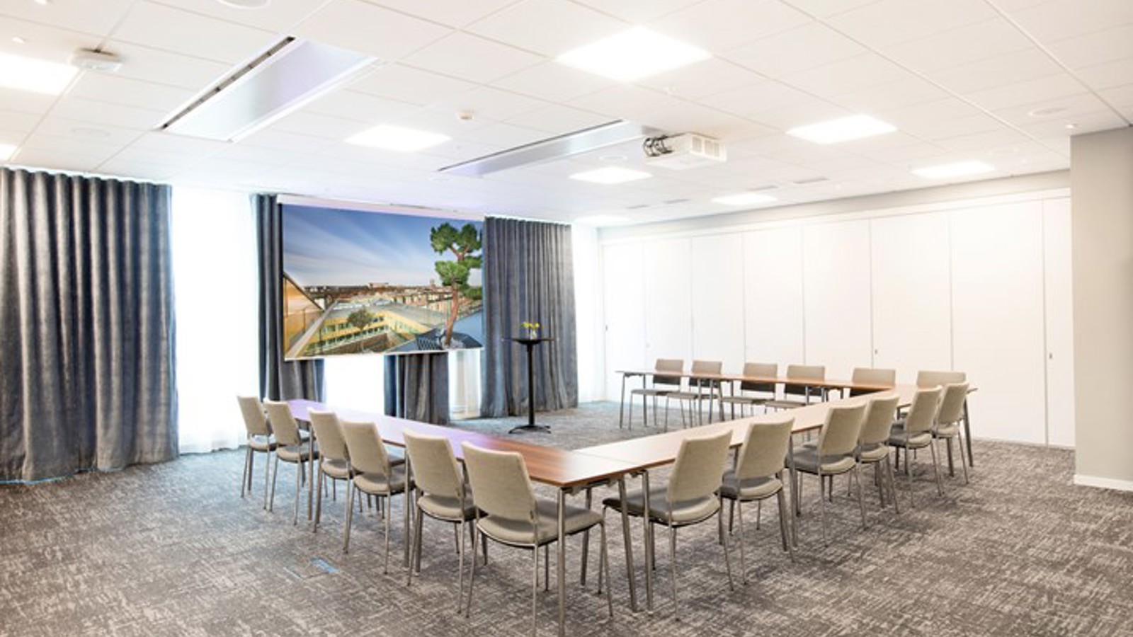 Konferensrum med u-sittning, grått golv, grå stolar och tända lampor i taket