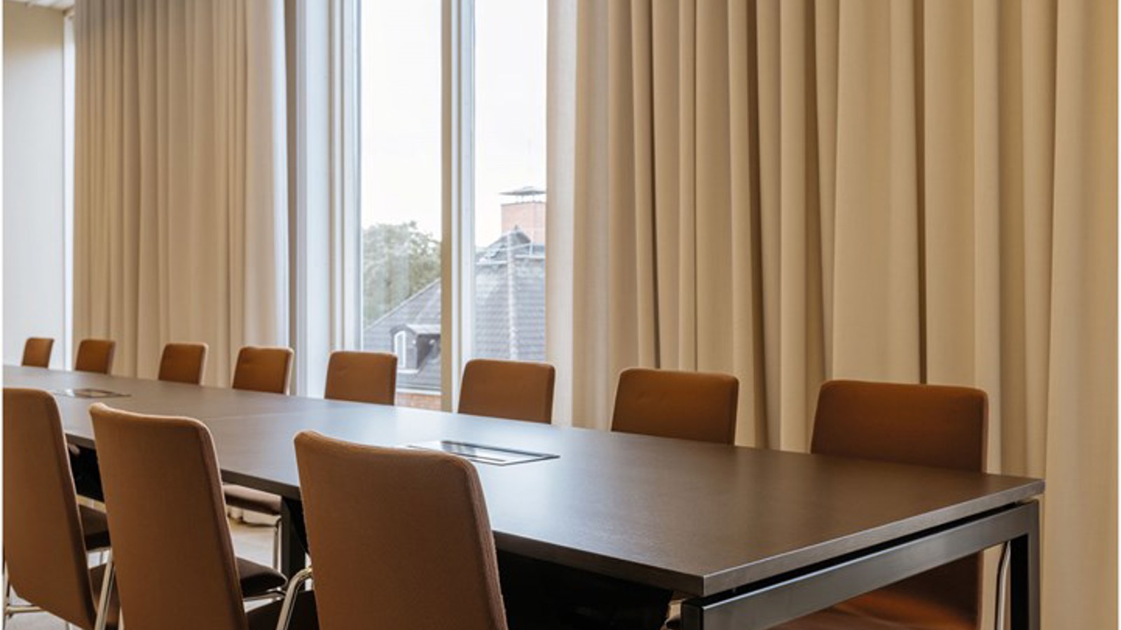 Konferensrum med styrelsesittning, brunt bord, bruna stolar, stora fönster och ljusa draperier