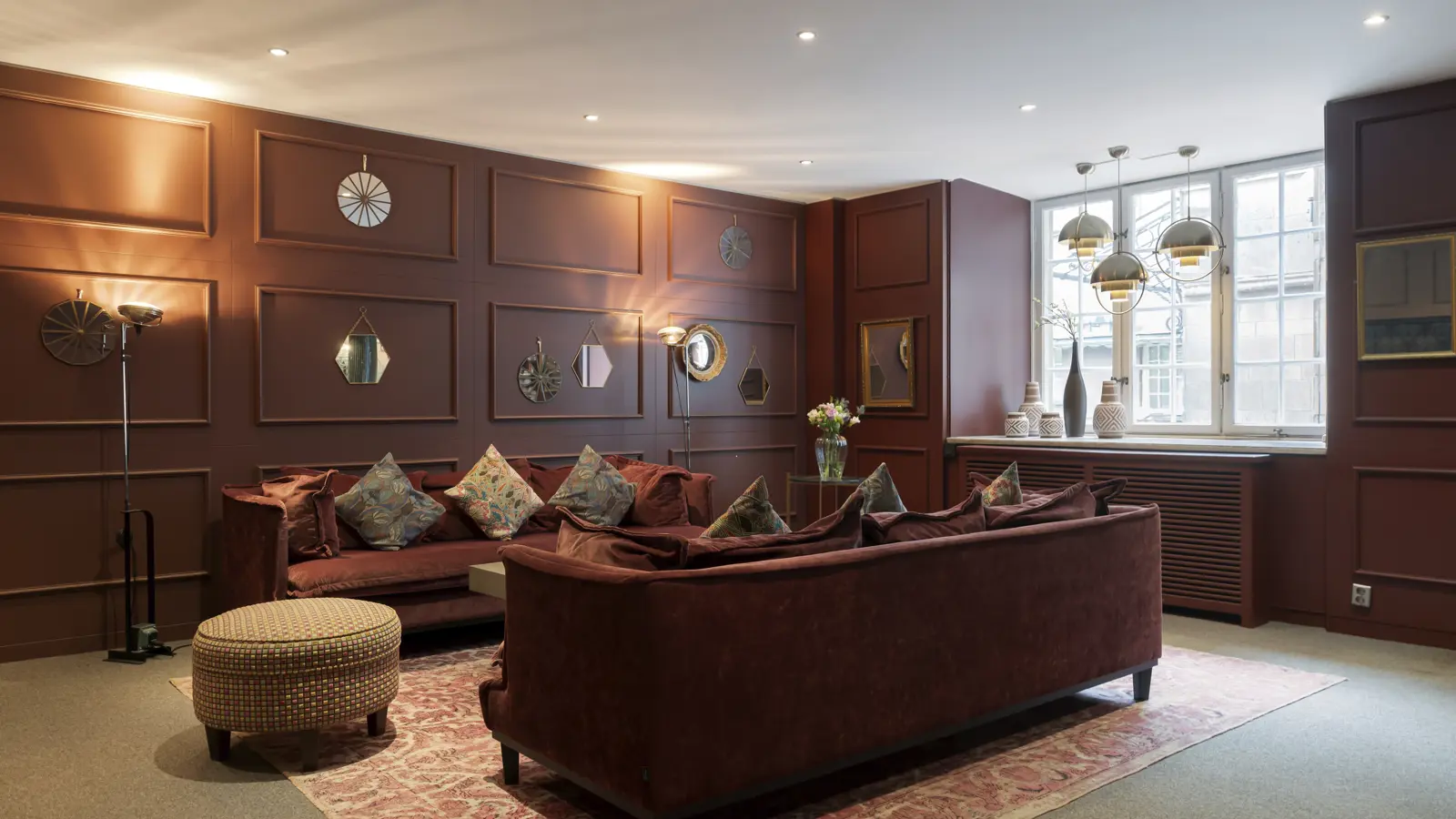 Elegant room with brown paneled walls, plush sofas, decorative mirrors, and modern lighting, featuring a large window overlooking a cityscape.