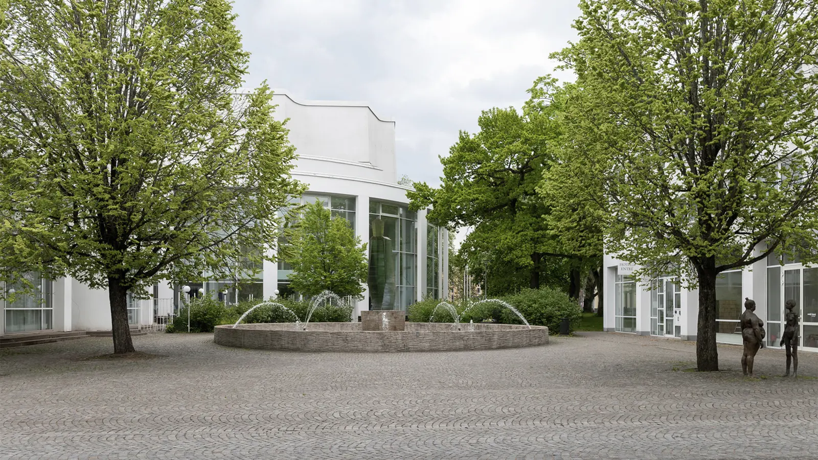 Modern building courtyard with a circular fountain in the center, surrounded by lush green trees and sculptures, set against a cloudy sky.