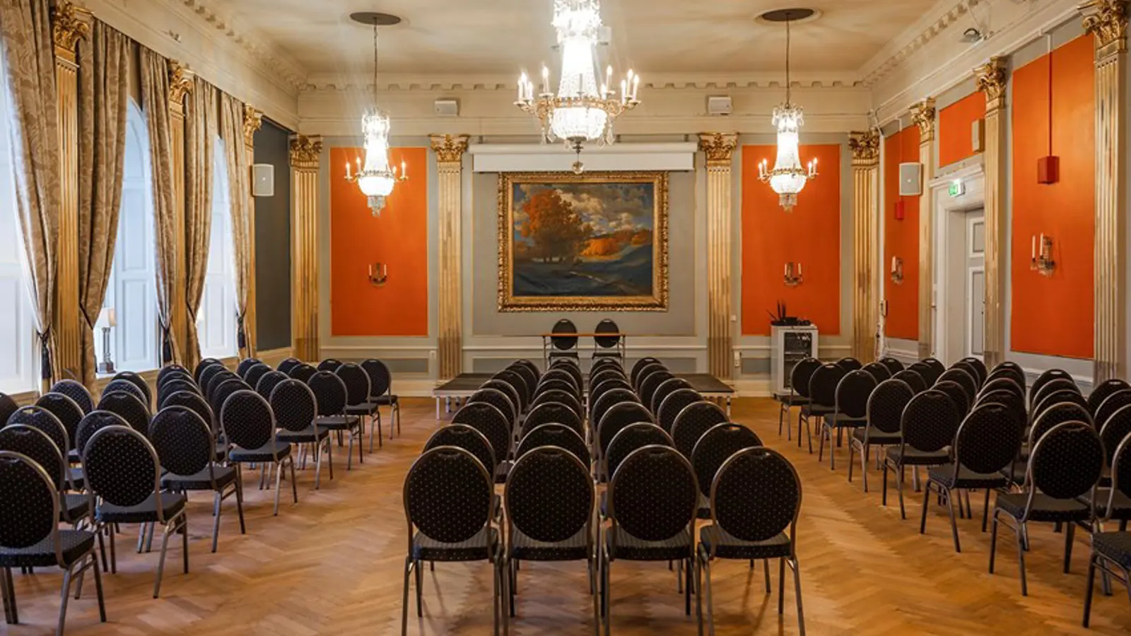 Large room with gold details, crystal chandelier, red walls and dark chairs lined up