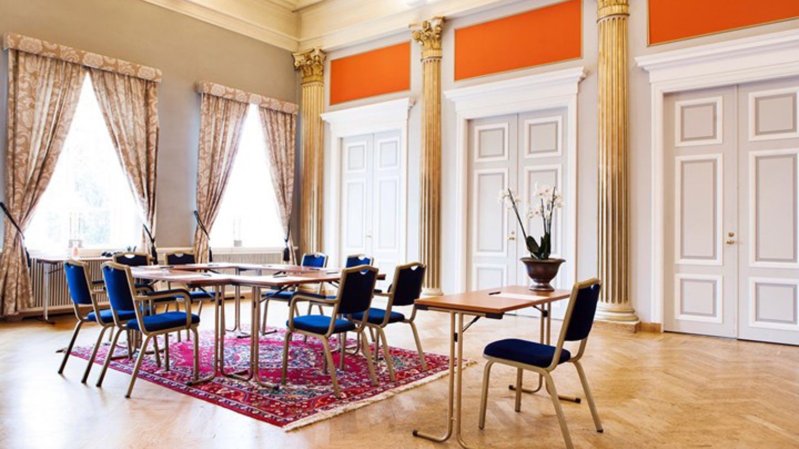 Konferensrum med högt till tak, guldpelare, stora fönster och blå stolar