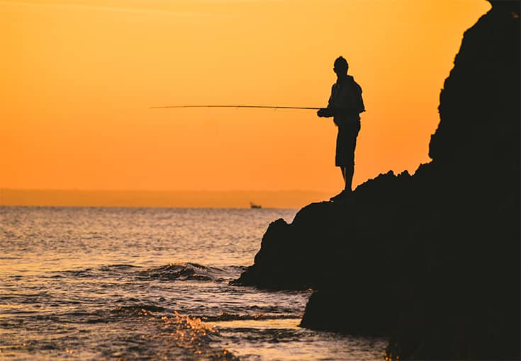 Man med fiskespö på en klippkant i solnedgången