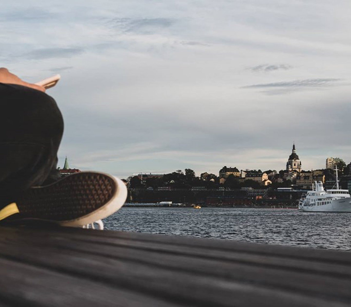 En person sitter på en brygga och håller en mobil med utsikt över Stockholm