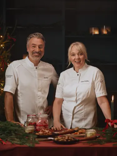 "Tareq Taylor and Carina Brydling, dressed in white chef’s jackets, stand by a Christmas-decorated table with candles, gingerbread biscuits and a Christmas tree in the background.