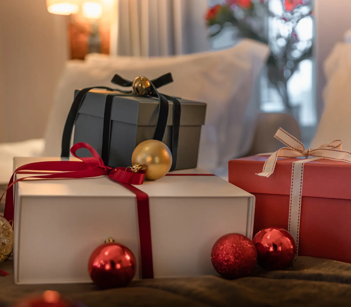 Holiday-themed scene featuring beautifully wrapped gift boxes adorned with ribbons and surrounded by festive ornaments, set on a cozy bed with soft lighting and a bouquet in the background.