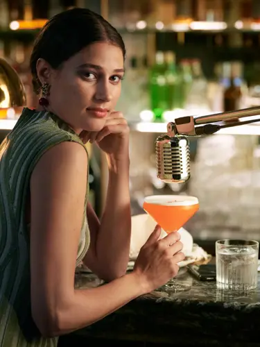 Kvinna i grön klänning som sitter vid bar med orange drink i handen och mikrofon över