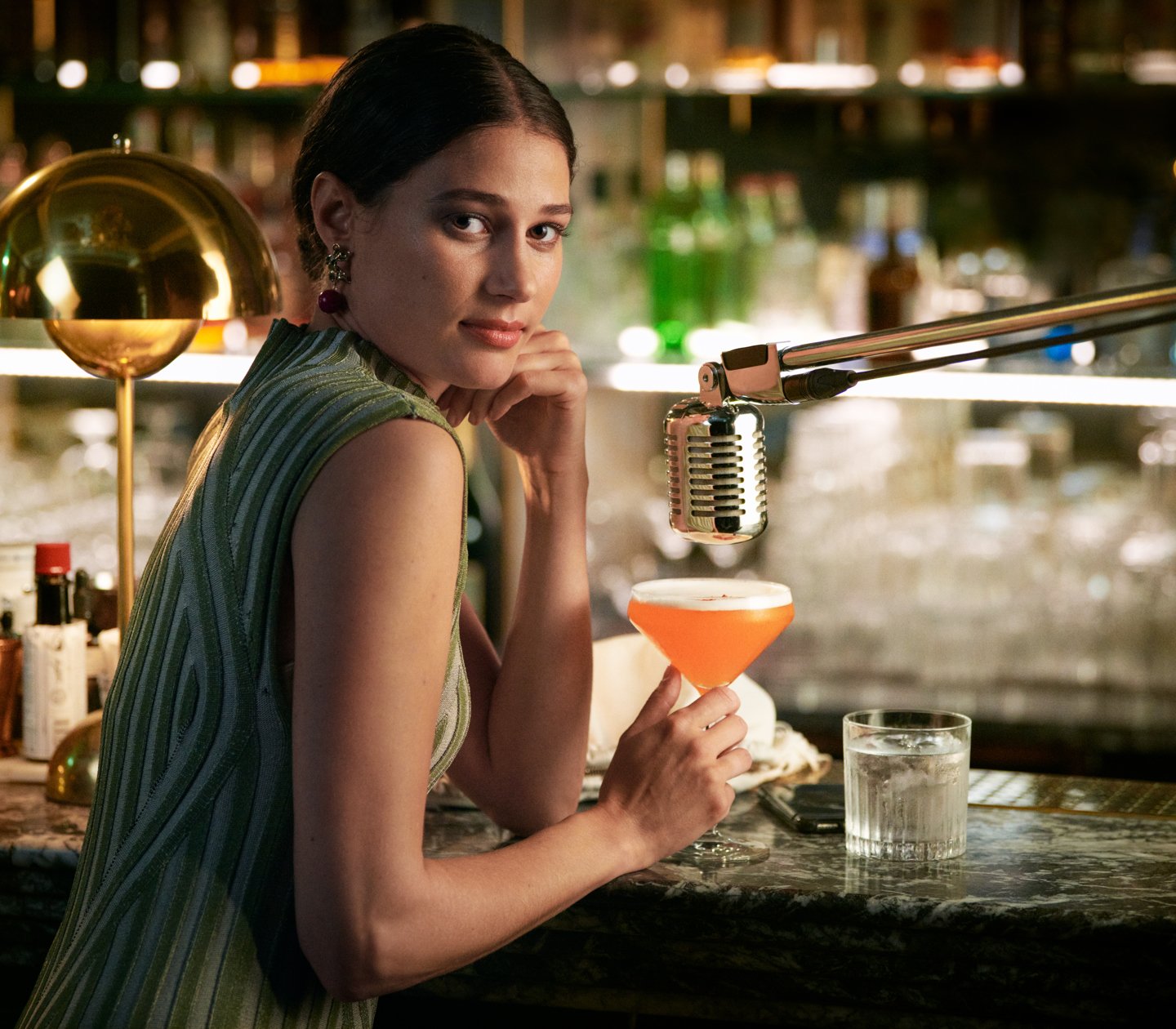 Woman in green dress sitting at bar with orange drink in hand and microphone over