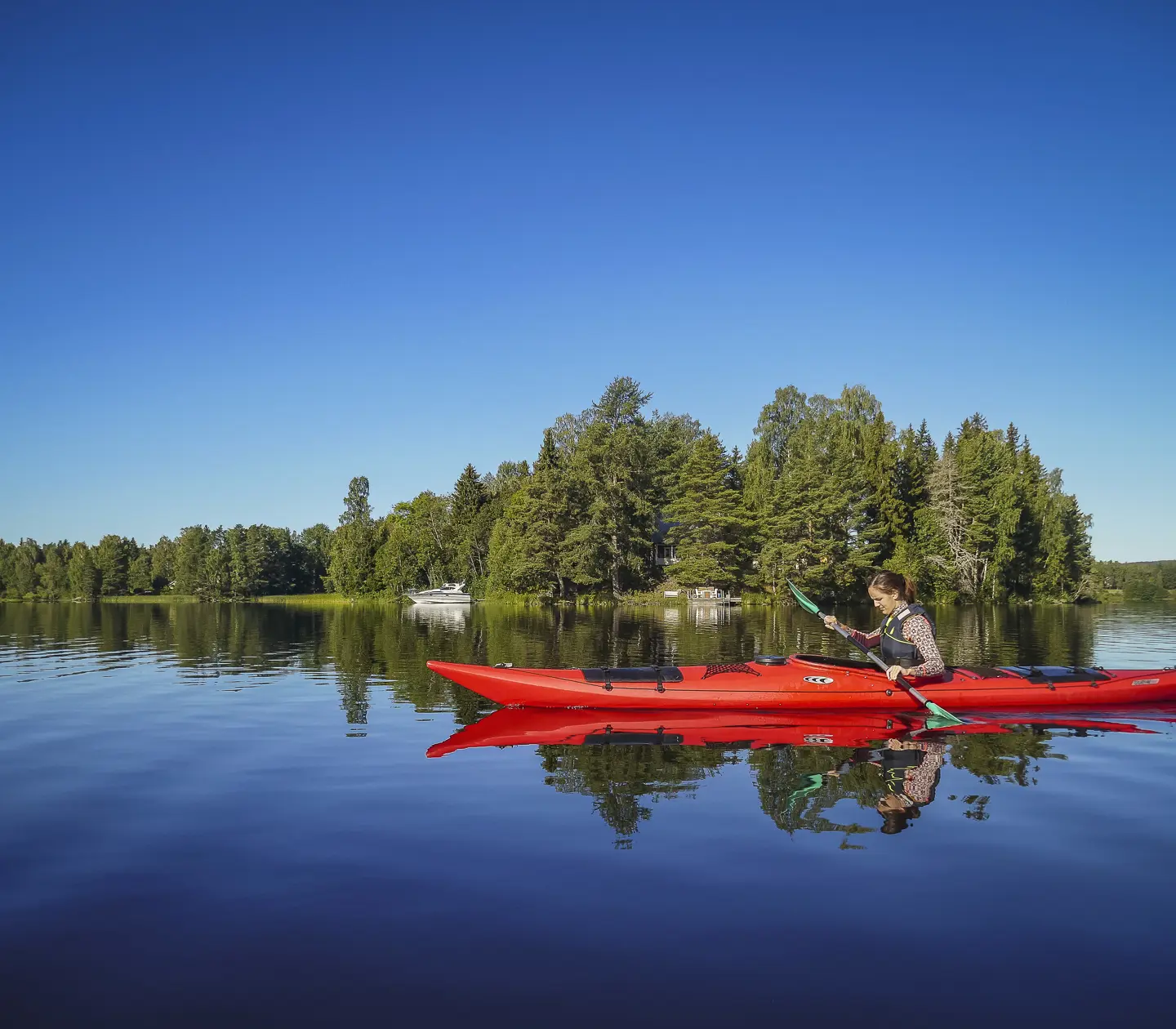 Person paddlar röd kajak på lugn sjö framför grönskande skog under klarblå himmel i Sverige
