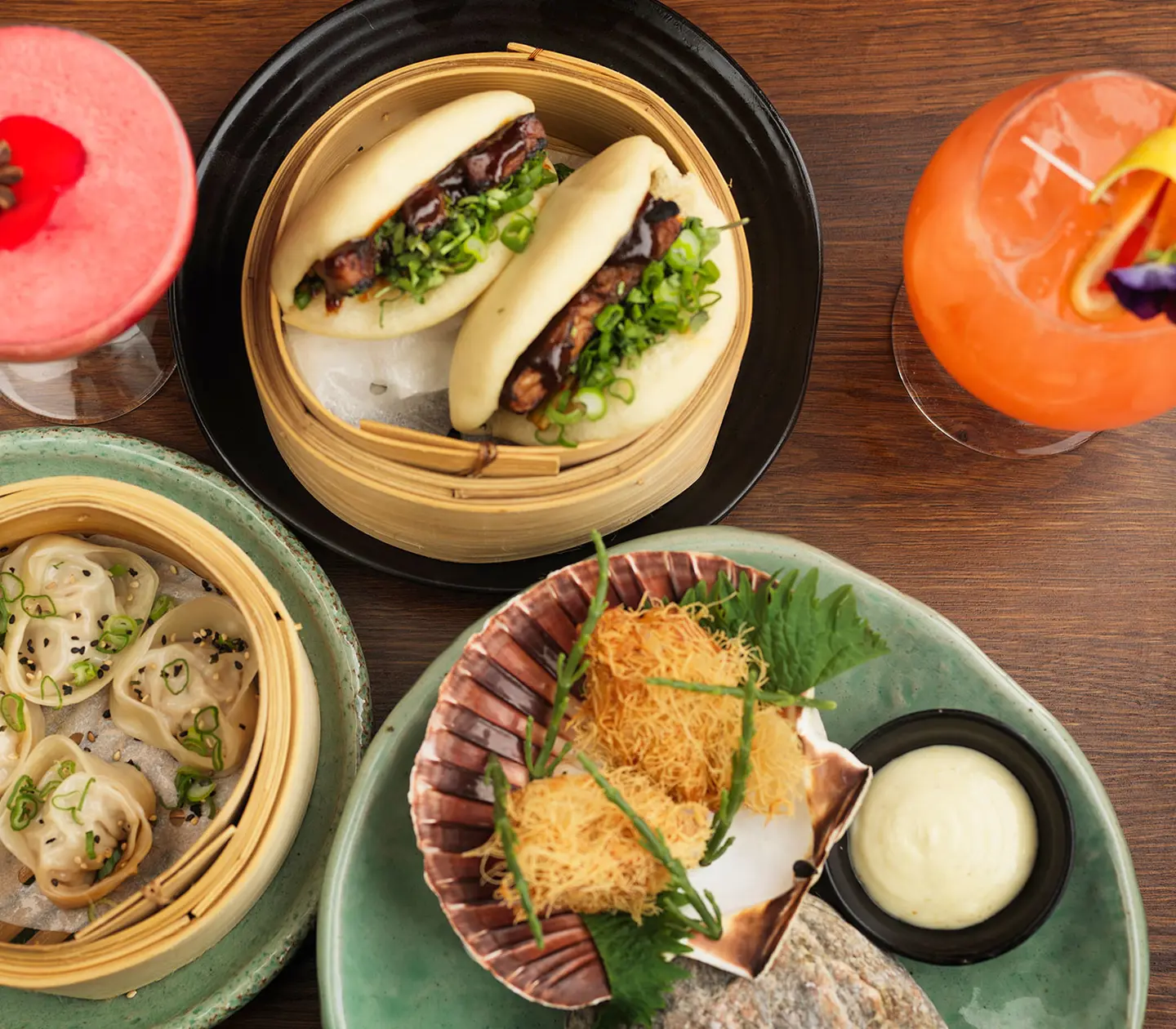 Asiatiska rätter med bao buns, dumplings och friterade skaldjur på ett bord, serverade med två färgglada cocktails