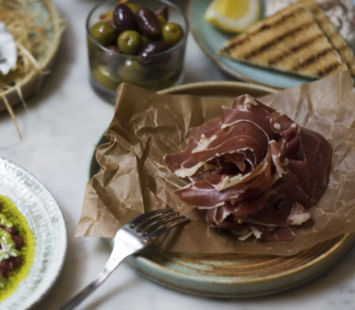Tunt skivad prosciutto på brunt papper, omgiven av tallrikar med gröna och svarta oliver, råbiff med dressing och grillat bröd, upplagt på ett marmorbord