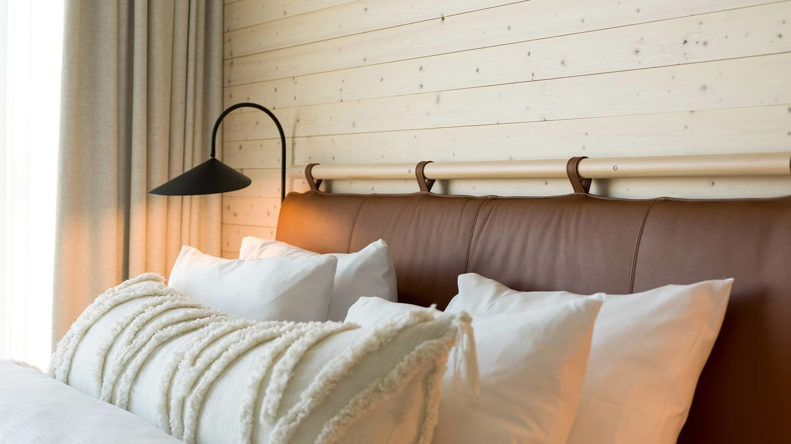 Fluffigt bäddad säng med sänggavel i brunt läder som står framför en vägg med ljus träpanel