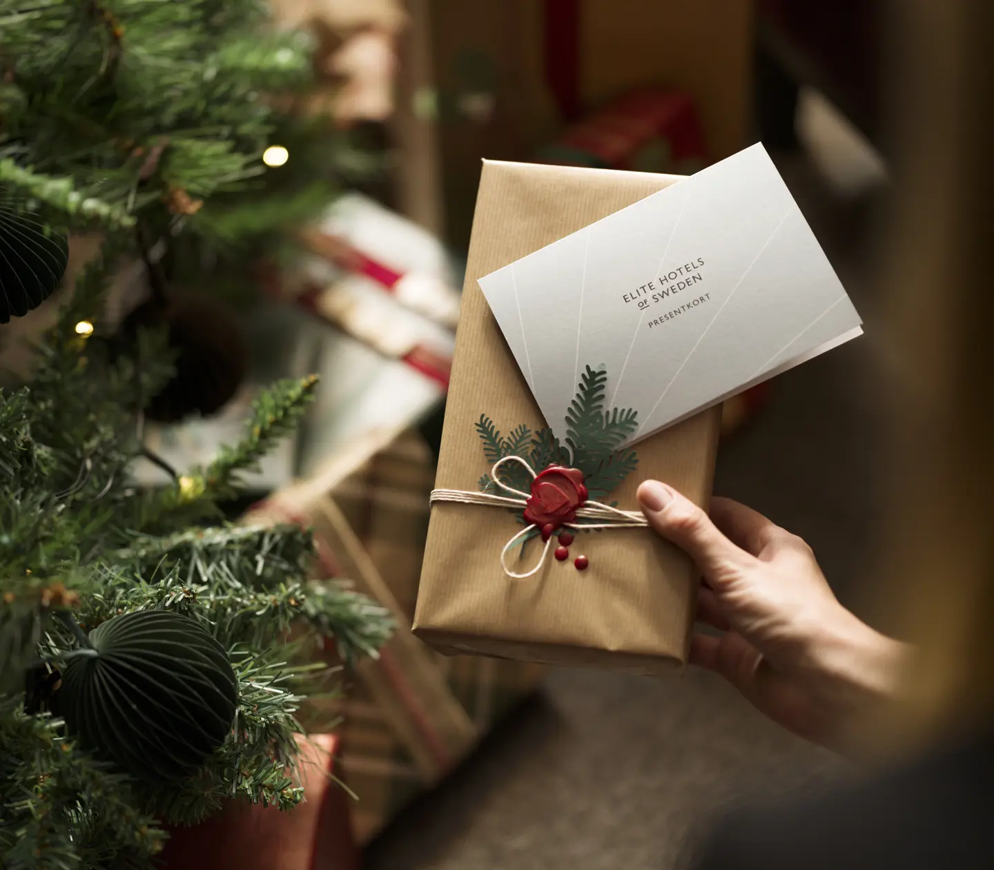 En julklapp med presentkort från Elite Hotels of Sweden och en julgran med julklappar i bakgrunden