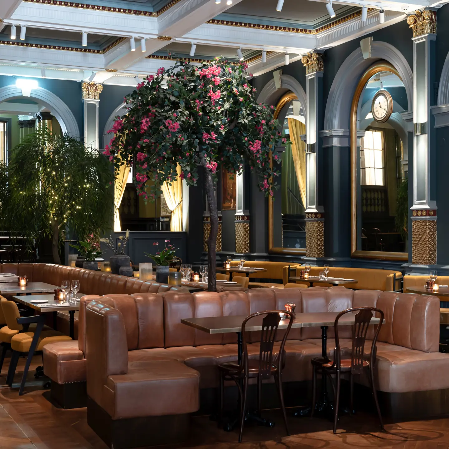 Eleganta interiör av en lyxig restaurang med stora bågformade speglar, bruna lädersoffor, klassiska trästolar, och en konstgjord blommande trädbeskärning i mitten, upplyst av mjukt ljus mot en mörk träkuliss.