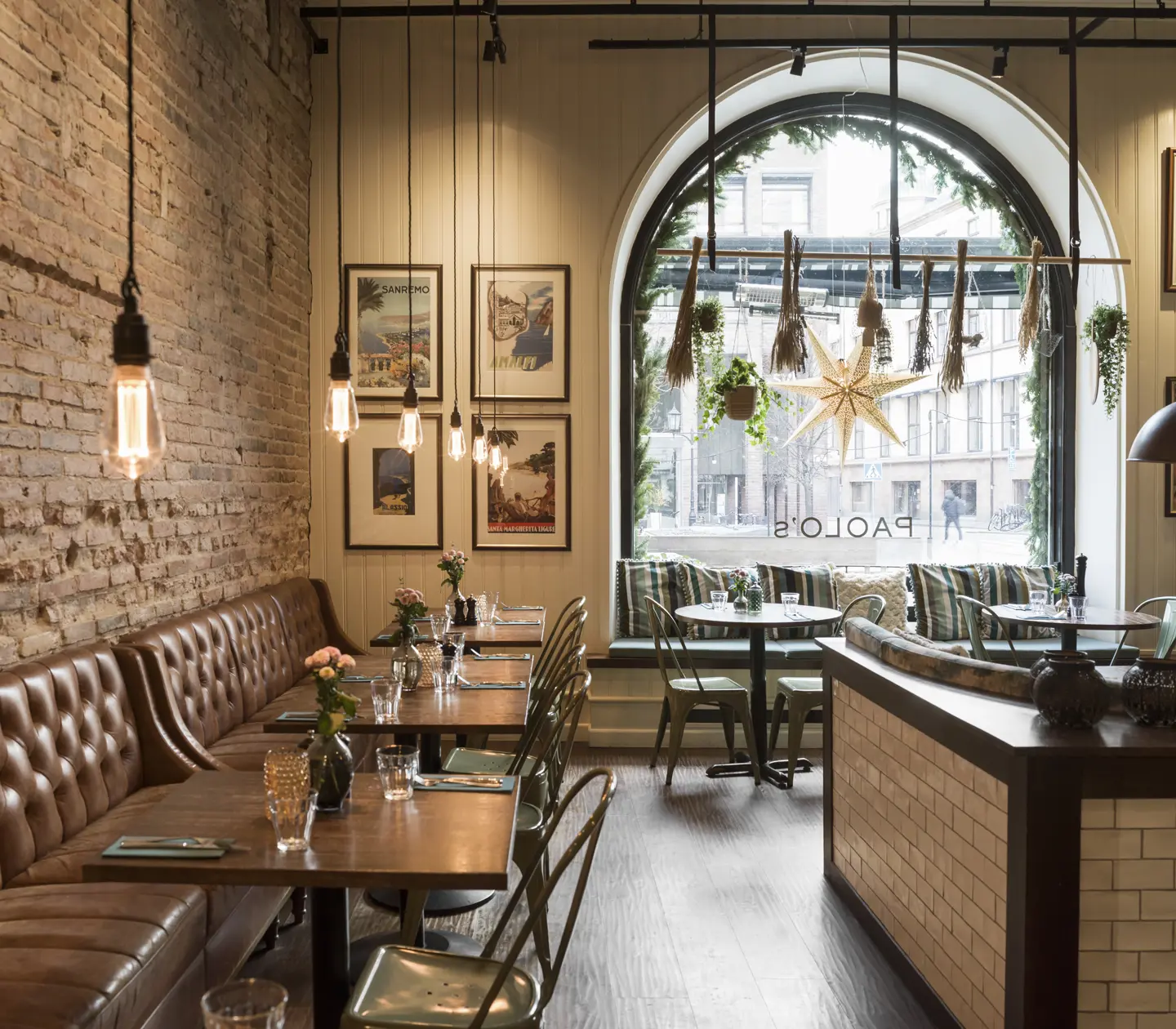 Modern restauranginteriör med tegelvägg, brunt läderbås och industriella ljuskällor. Stora fönster med utsikt över en stadsgata och dekorationer.