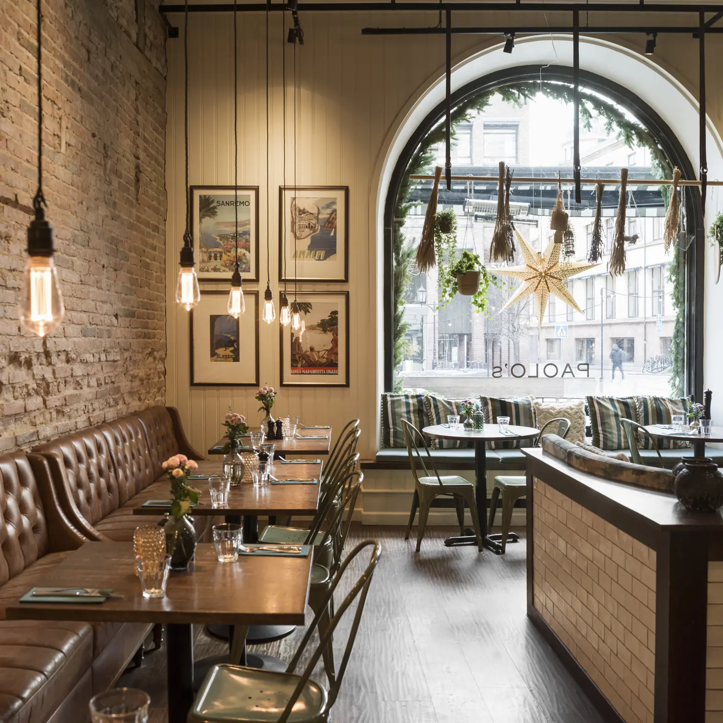 Modern restauranginteriör med tegelvägg, brunt läderbås och industriella ljuskällor. Stora fönster med utsikt över en stadsgata och dekorationer.