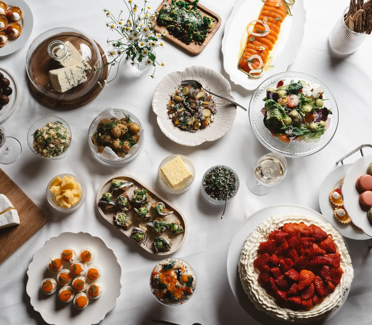 Svenskt smörgåsbord med olika delikatesser inklusive inlagd lax, sallad, bröd, ostar, tårta med jordgubbar, och macarons på ett uppdukat bord.