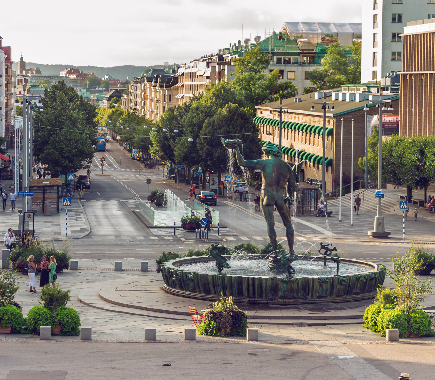 Utsikt över en livfull gata och torg i Göteborg med människor som promenerar, staty och fontän i förgrunden, och stadsmiljö i bakgrunden.